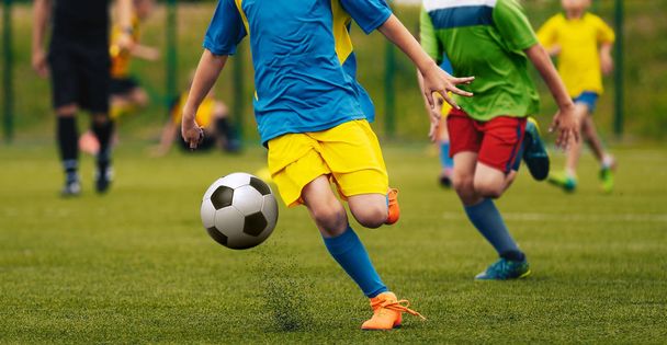 Αγόρια στο μπλε, κίτρινο και πράσινο μπλούζες ζέρσεϊ κλοτσιές ποδόσφαιρο στο γήπεδο ποδοσφαίρου. Ποδόσφαιρο παίκτες τρέχουν σε δράση. Σχολείο νεότητα Ποδοσφαιρικό αγώνα. Διαιτητής ποδοσφαίρου στο παρασκήνιο. - Φωτογραφία, εικόνα