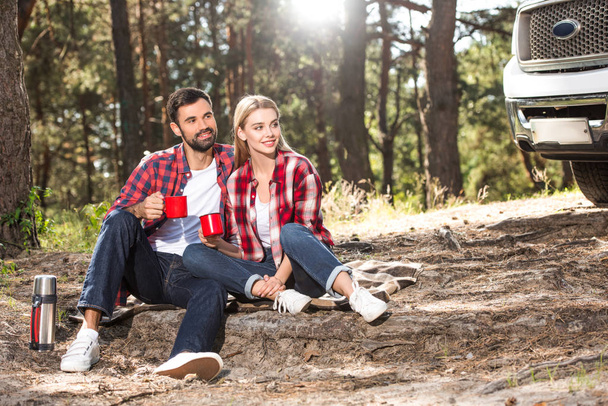happy young couple sitting on ground with coffee cups near car outdoors  - Photo, Image
