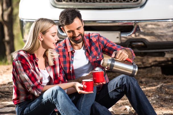 Gelukkig jonge man gieten koffie naar vriendin cup van thermos in de buurt van de auto buiten  - Foto, afbeelding