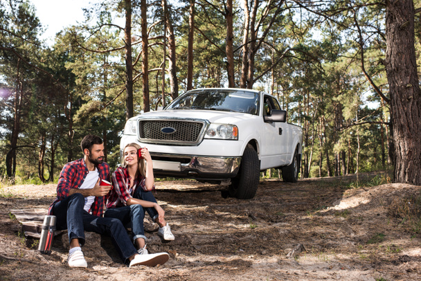 young couple sitting on ground with coffee cups near pick up car in forest - Photo, Image