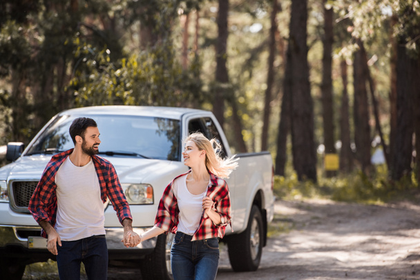 beautiful cheerful couple holding hands and running in forest with pickup truck - Фото, зображення