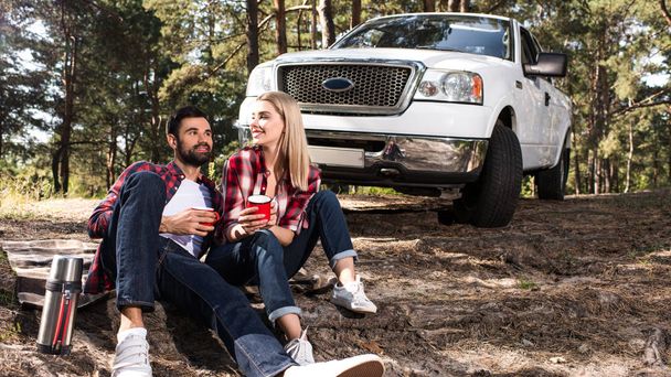 sonriente joven pareja sentada en el suelo con tazas de café cerca recoger coche en el bosque
  - Foto, Imagen