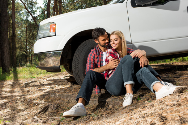 young couple sitting on ground and embracing near pick up car outdoors  - Zdjęcie, obraz