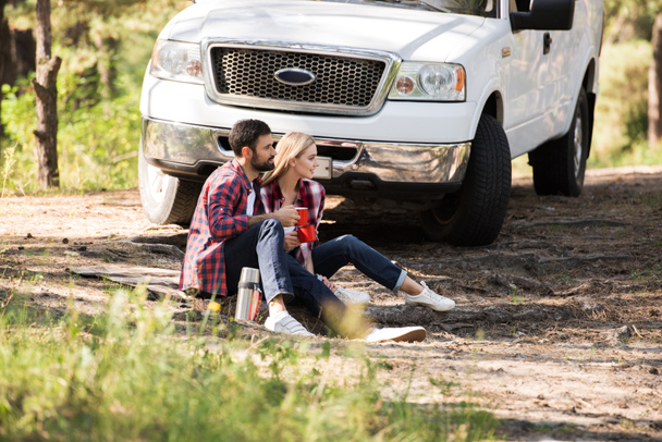 пара сидить на пікніку з термосом в лісі з машиною позаду
 - Фото, зображення