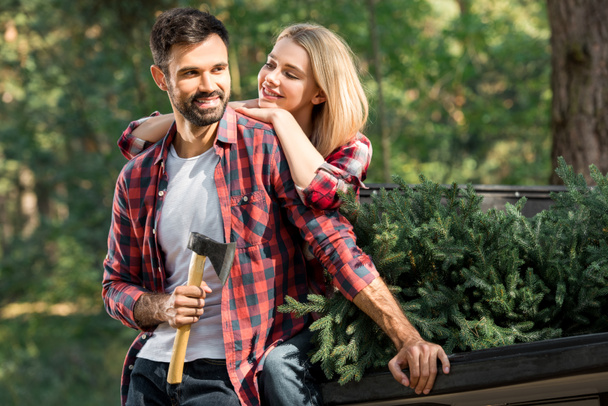 cheerful young man holding axe and standing near girlfriend sitting in car trunk with christmas tree in forest - Foto, immagini