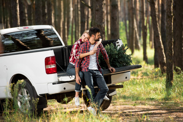foco seletivo de bela jovem mulher abraçando namorado por trás enquanto sentado no porta-malas do carro com árvore de natal na floresta
 - Foto, Imagem