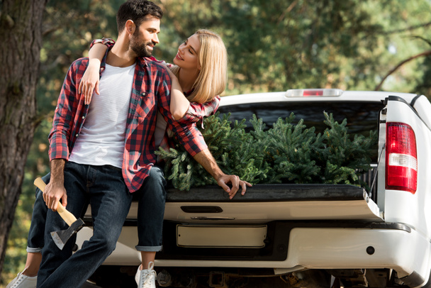 handsome young man holding axe and standing near girlfriend sitting in car trunk with christmas tree in forest - 写真・画像
