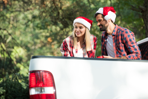 jeune couple souriant dans des chapeaux de Père Noël avec des tasses assis dans le coffre de la voiture dans la forêt
  - Photo, image