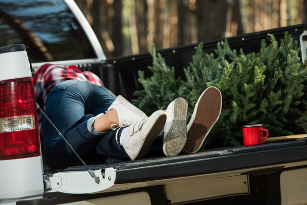 partial view of young couple laying in car trunk with christmas tree and cup outdoors  - Photo, Image