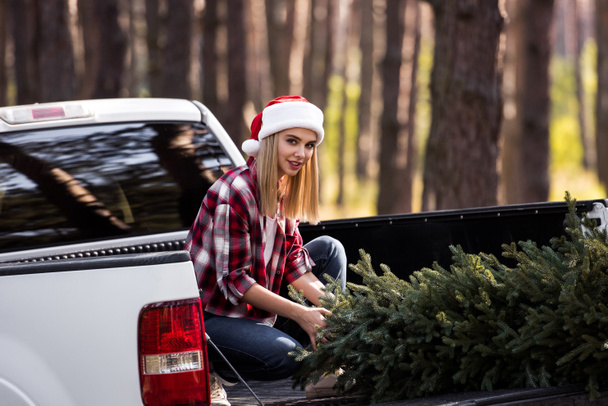glückliches Mädchen mit Weihnachtsmütze sitzt auf Pickup-Truck mit Tannenbaum für Weihnachten - Foto, Bild