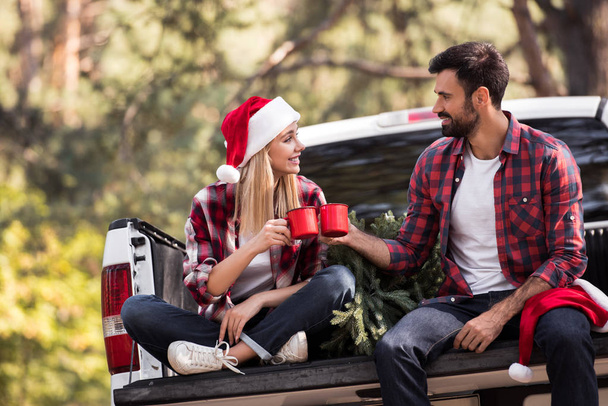 junges Paar in Weihnachtsmannmützen klingelt mit roten Tassen, während es auf einem Pickup-Truck mit Weihnachtsbaum sitzt  - Foto, Bild