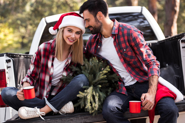 όμορφο ζευγάρι σε καπέλα santa κρατώντας κόκκινα φλιτζάνια ενώ κάθεται σε φορτηγό επαναλείψεων με έλατο για τα Χριστούγεννα  - Φωτογραφία, εικόνα