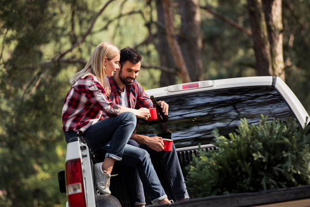 junges Paar gießt Wein in Becher, während es auf Pickup-Truck mit Weihnachtsbaum im Wald sitzt - Foto, Bild