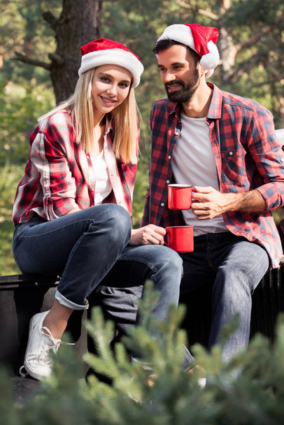 smiling couple in santa hats holding red cups while sitting on pickup truck with fir tree for christmas in forest - Photo, Image