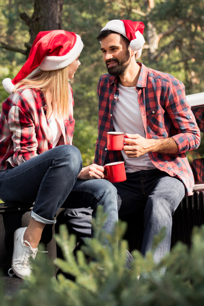 happy couple in santa hats holding red cups while sitting on pickup truck with fir tree for christmas in forest - Φωτογραφία, εικόνα