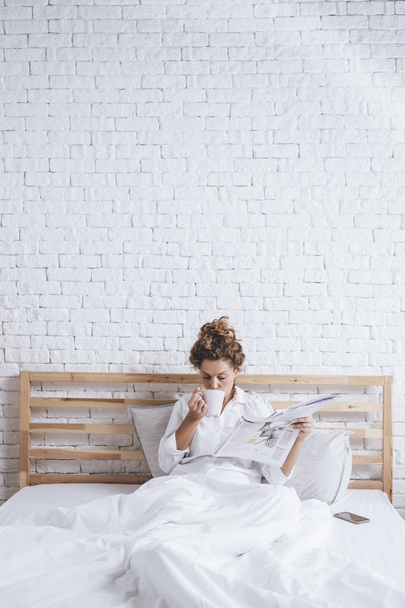 Pretty Caucasian woman reading newspapers and drinking coffe in bed - Foto, imagen