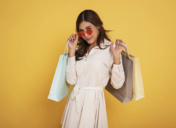 Портрет збудженої красивої азіатської дівчини в сукні та сонцезахисних окулярах, що тримає сумки для покупок ізольовані на жовтому тлі
. - Фото, зображення