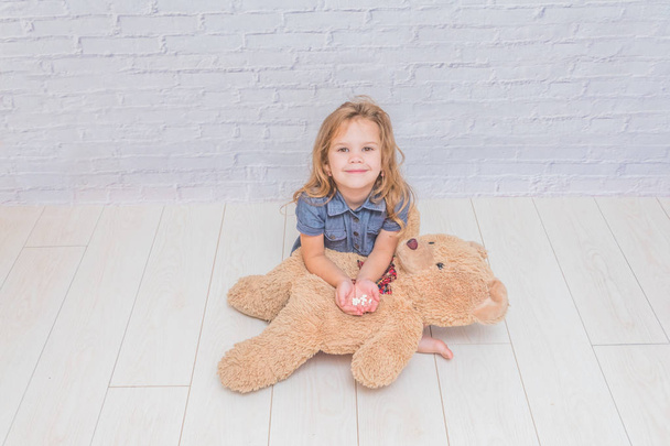 the girl, child treats a toy bear, a pill in front of a white brick wall - Foto, afbeelding