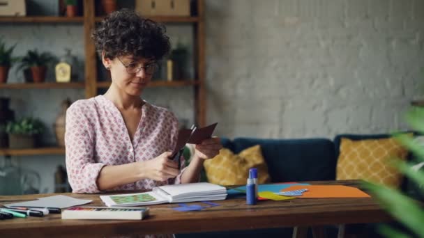 Creative young woman is cutting piece of paper with scissors creating collage then putting it in notebook and checking design. Creativity, workspace and art concept. - Filmagem, Vídeo