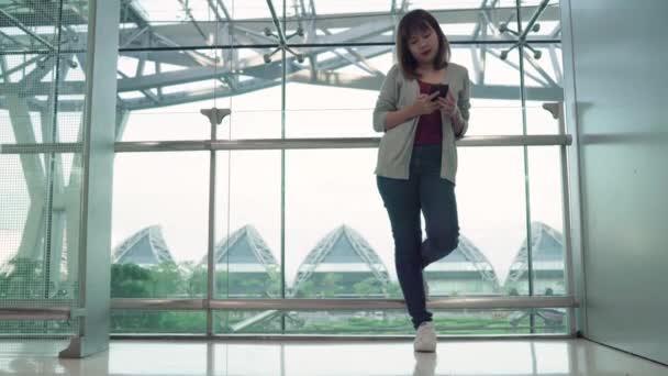Attractive casual happy young Asian woman in international airport, using and checking her smartphone standing in terminal hall while waiting for her flight to board airplane at the departure gate. - Footage, Video