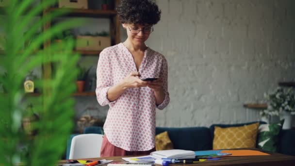 Smiling curly-haired lady is taking pictures of desk creating flat lay using smartphone camera standing in modern apartment. Modern technology and design concept. - Filmagem, Vídeo