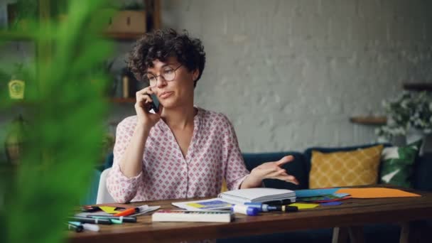 Pretty young woman is glasses is talking on mobile phone and smiling sitting at desk with stationery at home. Communication, houses and technology concept. - Metraje, vídeo