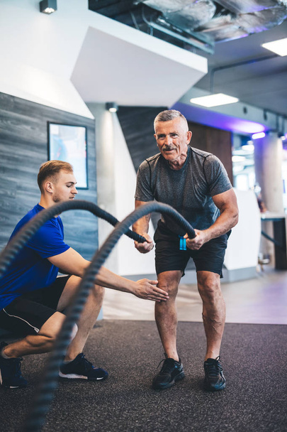 Homme âgé faisant de l'exercice avec des cordes à la salle de gym. Formation personnelle. Mode de vie sain
. - Photo, image