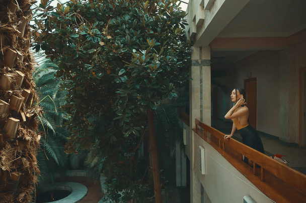 Pretty sensual young woman standing in hotel balcony, looking out enjoying a vacation surrounded by exotic trees. Recreation and relaxation concept. - Photo, Image
