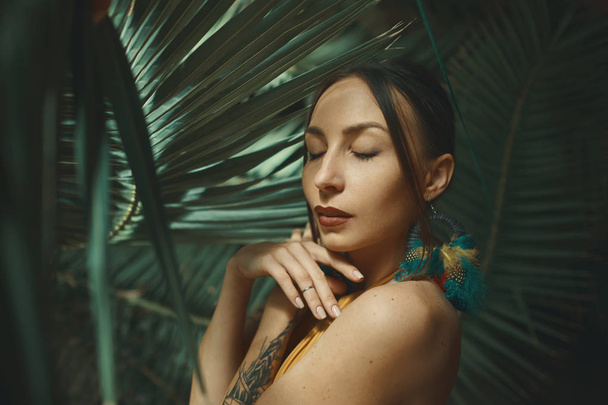 Beauty Woman. Beautiful Young Exotic Dreamy Female, Closed Eyes, touching Her Skin. Portrait on Palm Leaves Background. Healthcare. Perfect Skin. Face. Big Circular Earrings with Colorful Feathers. - Foto, immagini