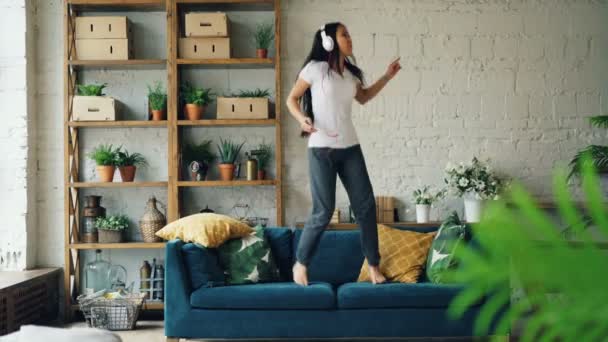 Attractive Asian woman is having fun at home listening to music through headphones holding smartphone, singing and dancing jumping on sofa. Modern technology and people concept. - Filmati, video