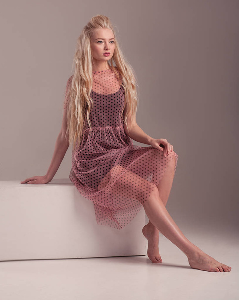 Blonde barefoot model in transparent dress is posing in studio on pink background. - Photo, image