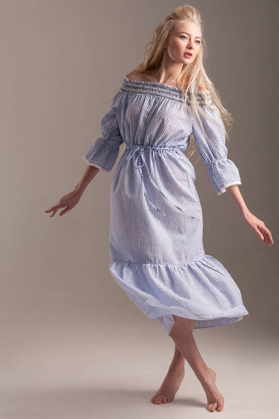 Blonde barefoot model in blauwe jurk is poseren in studio op grijze achtergrond. - Foto, afbeelding