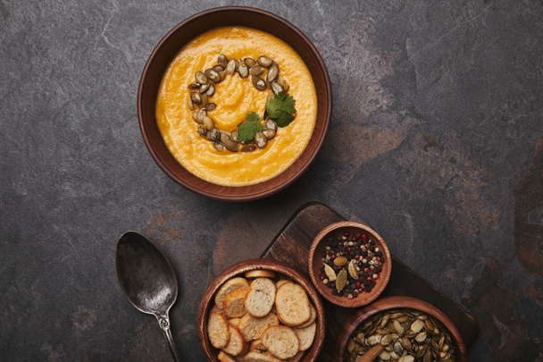 bowl with tasty pumpkin soup, spices, rusks, pumpkin seeds and spoon on dark surface  - Photo, Image