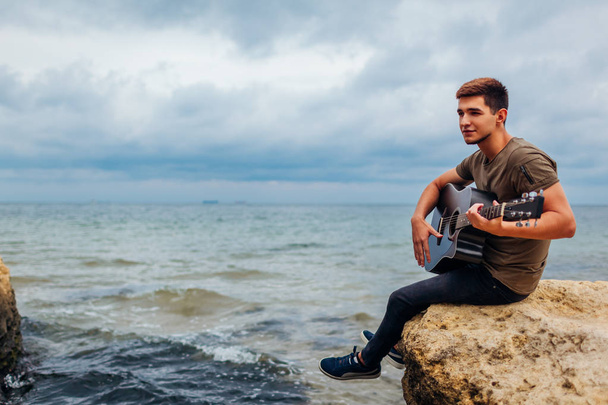 Young man with acoustic guitar playing on beach surrounded with rocks on rainy day. Guy enjoying hobby - 写真・画像