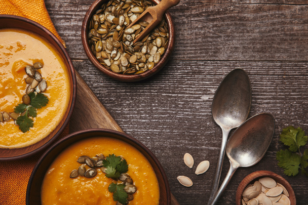 вид вкусного здорового тыквенного супа в мисках, ложках и семенах тыквы на деревянном столе
 - Фото, изображение