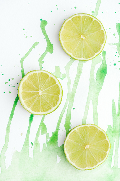 elevated view of three pieces of ripe limes on white surface with green watercolor - Foto, imagen