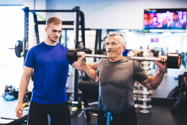 Older man and personal trainer at the gym. Lifting weights. Indoor workout. - Foto, Imagem