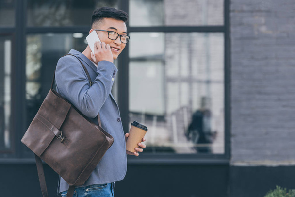 handsome asian man with leather backpack and coffee to go talking on smartphone  - Foto, immagini