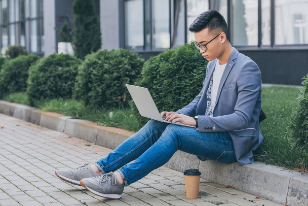 confident asian freelancer working on laptop while sitting on sidewalk with disposable cup of coffee - Фото, изображение