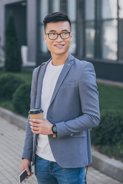 sorridente uomo asiatico in giacca grigia in possesso di caffè per andare e smartphone
 - Foto, immagini
