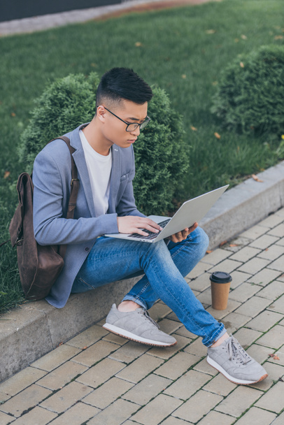 красивий азіатський чоловік працює з ноутбуком, сидячи на тротуарі
 - Фото, зображення