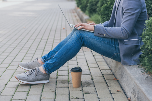 cropped view of freelancer with coffee to go working on laptop while sitting on sidewalk - Photo, Image