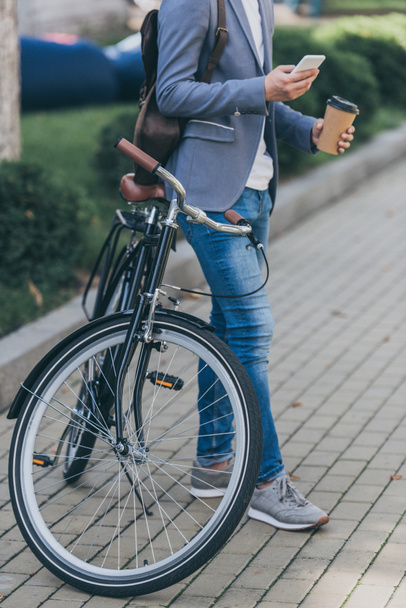 vista recortada del hombre con café para ir con el teléfono inteligente y apoyado en la bicicleta
 - Foto, Imagen