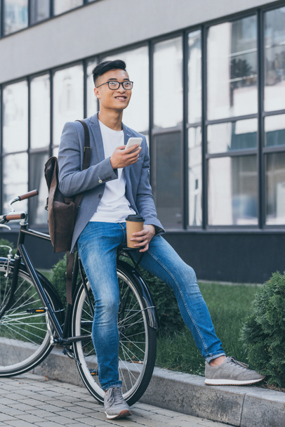 hombre asiático feliz con taza de café desechable usando teléfono inteligente y sentado en bicicleta
 - Foto, Imagen