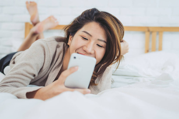 Mulheres asiáticas felizes estão usando telefone inteligente na cama de manhã. Mulher asiática na cama verificando aplicativos sociais com smartphone. Mulher sorridente surfando rede com celular em casa. Conceito de viciado móvel
.  - Foto, Imagem