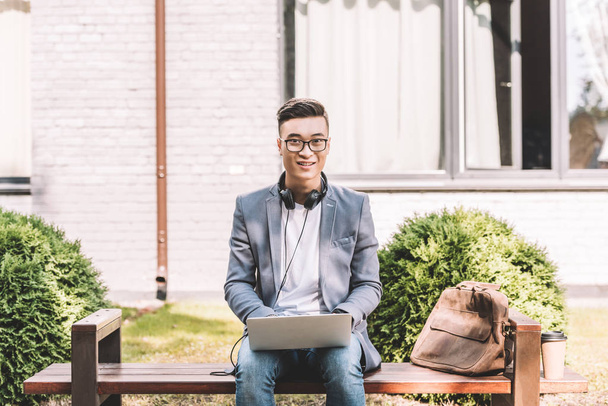 sorridente asiatico freelancer working su laptop mentre seduta su panca con borsa in pelle
 - Foto, immagini