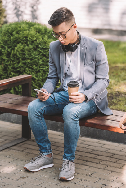 stylish asian man with coffee to go, smartphone and headphones sitting on bench - 写真・画像