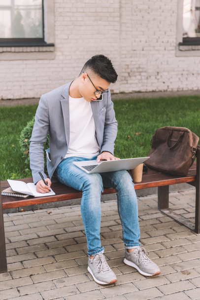 Aziatische freelancer schrijven in planner terwijl praten op smartphone en werken op de laptop op de Bank - Foto, afbeelding