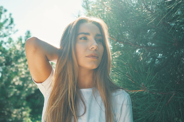 Close-up young woman with beautiful face features touching hair with hand smiles enjoying life with sun rays on her face with pine in background. Light filters used. - Φωτογραφία, εικόνα