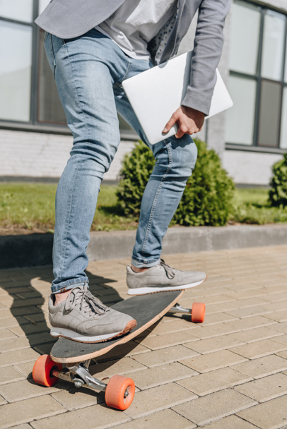 cropped view of stylish man with laptop skateboarding on longboard - Фото, изображение